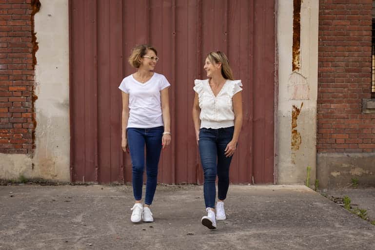 élène et Séverine, l’équipe de pas.prasse en promenade Hélène et Séverine, souriantes, marchent côte à côte devant un portail en briques rouges, illustrant leur complicité et leur dynamisme.