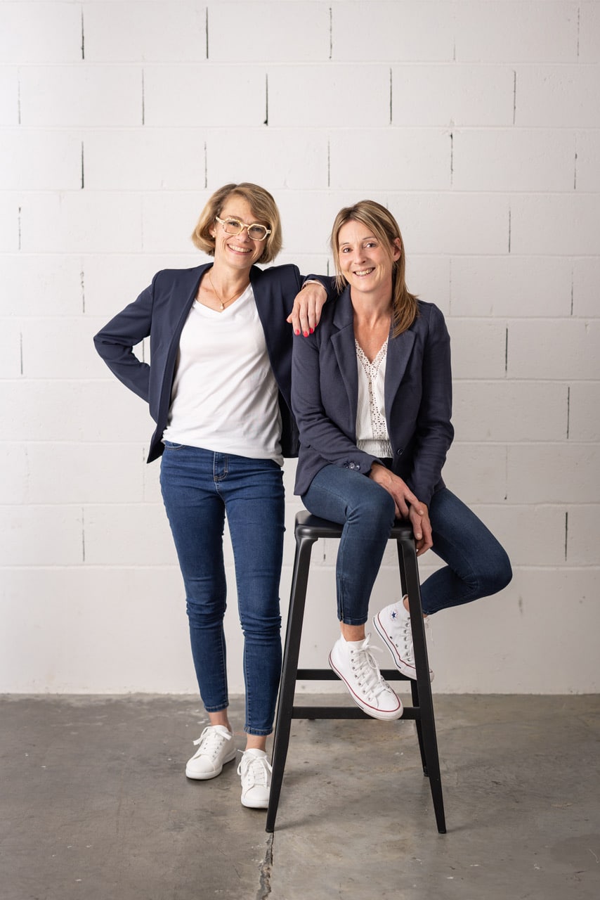 Hélène et Séverine, l’équipe de pas.prasse Hélène et Séverine, souriantes, vêtues de vestes et de jeans, posent côte à côte devant un mur blanc. Elles représentent l’équipe de pas.prasse dans une ambiance à la fois décontractée et professionnelle.