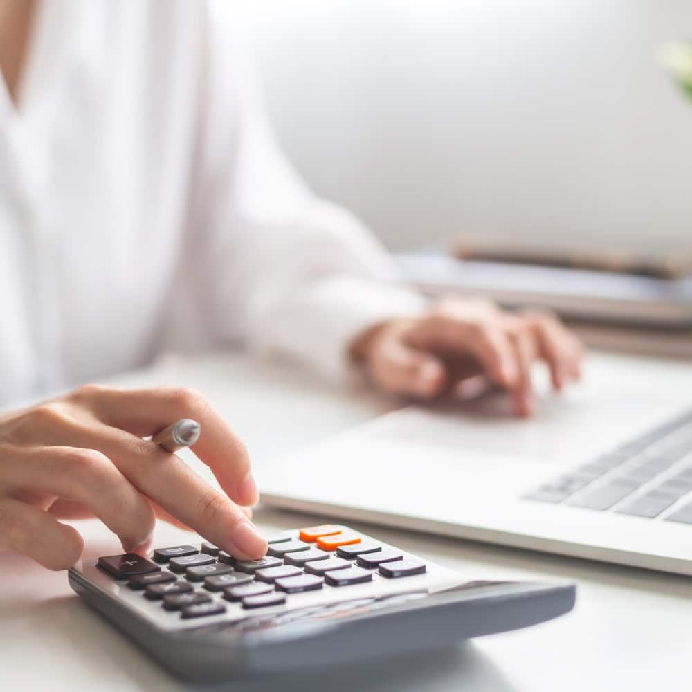 Gestion administrative commerciale Gros plan sur les mains d’une personne en chemise blanche utilisant simultanément une calculatrice et un ordinateur portable, illustrant un travail de comptabilité ou de gestion administrative.
